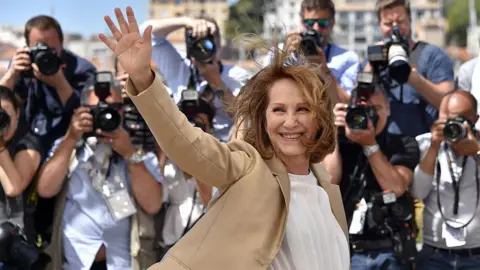Nathalie Baye, a woman with short brown hair, waves in front of a crowd of photographers in Cannes. She wears a camel-coloured blazer and a white t-shirt. She is smiling.