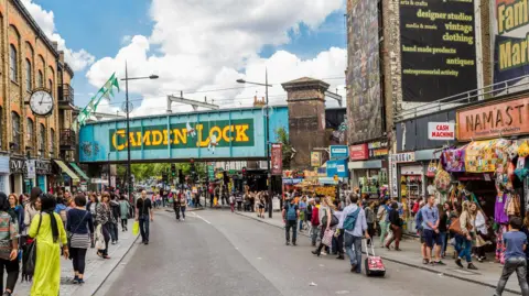 Crowds of people walk on pavement on either side of a road. The road goes underneath a blue railway bridge in the distance that has the words Camden Lock painted on it in bright yellow capital letters. There are buildings, market-style shop fronts and signage all around. 