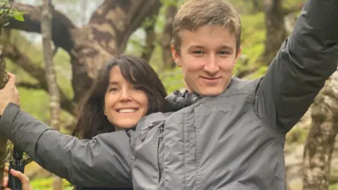 Family photo James, smiling wearing a grey coat with his arms holding on to the branches of a tree. His mother, with long brown hair, is standing behind him with her head resting on his shoulder.