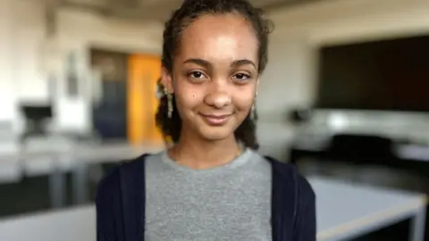 Martin Giles/BBC Iona is standing in a classroom. She has long dark hair, swept back from her forehead. She is wearing earrings and is smiling at the camera.