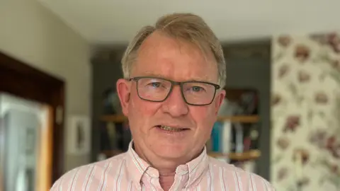 BBC Dr Kieran McManus has short brown hair and is wearing black square glasses with a pink striped shirt. Behind him is a bookcase and a white floral pattern wall.
