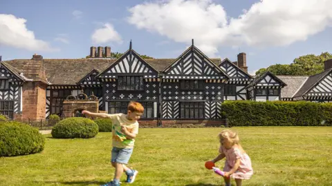 National Trust Images/James Beck A young boy with red hair, yellow t-shirt and blue denim shorts plays on the lawn in front of Speke Hall alongside a young girl in a pink dress with blond hair holding a ball on a clear day.