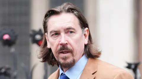 PA Media Richard Hall wears a brown blazer and blue shirt and tie outside court. He has medium length brown hair and a brown moustache and beard.