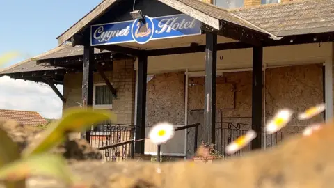 A shot of the wooden archway of the entrance of the hotel. There is a wooden archway with a blue sign in the middle reading "cygnet hotel" with an image of a swan between the words. Daisies and leaves are out of focus in the foreground. 
