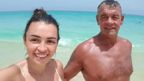 Abbey Wright A young white female with brown hair pictured with a middle aged man (her Dad) with grey hair on a beach in front of the sea. 