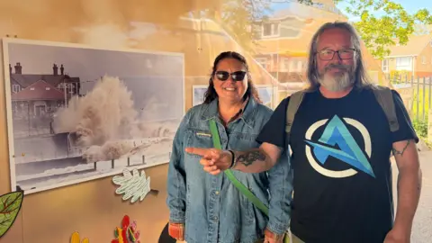 JasmineLowe/BBC News A lady wearing a denim jacket, with curly hair and sunglasses on under a viaduct next to an image on the wall showing waves crashing into the pier. Her husband beside her has long grey hair and is wearing a black top and a back pack. He's pointing to the image with the waves on as he said it's his favourite. 