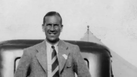 Essex Cricket Club A black and white image of Laurie Eastman. He has short hair and is wearing a tweedy jacket over a white shirt and stripped tie. He is smiling, leaning on a just glimpsed car.