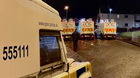a police officer stands between three psni landrovers in the creggan area of derry at night. Debris can be seen on the road