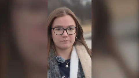 A young woman with glasses and long brown hair looks at the camera
