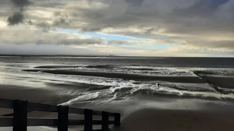 Weather Watchers/The Cleeve Rambler A moody photography of wet sand at a beach which appears almost grey in colour. The tide appears to be going out with tide lines visible on the sand. The brown water from the Severn is visible out towards the horizon. The sky is cloudy with a small patch of blue in the centre of the frame. A railing along a jetty going down to the beach is visible in the left of the shot.