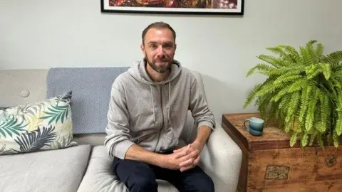 Sam with short dark hair and a beard, wearing a grey hooded top, sitting on a sofa beneath a framed picture on the wall.