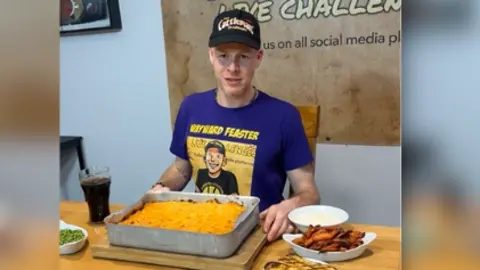 A still from a video, a man sits behind a massive cottage pie in a tray with side plates full of food beside him. He wears a cap and t-shirt which says 'Wayward Feaster'