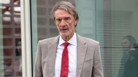 Sir Jim Ratcliffe, who has brown greying hair and a grey stubble beard, wears a beige linen suit jacket with a white shirt and red tie