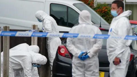 PA Media Forensics officers are at the scene of the shooting on Kingsheath Avenue where nine-year-old Olivia Pratt-Korbel was fatally shot. Two forensic officers are standing near the police cordon while another three are looking on the ground. They are all wearing white overalls and masks.