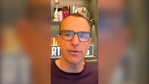 Martin Lewis wears glasses and a purple top. He sits in front of bookshelves