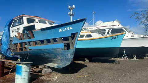 BBC Damaged boat in dry dock with the name Aliva painted on the side, next to another repaired boat with the name Joyce on the side. The boats are blue and white against a bright blue sky.