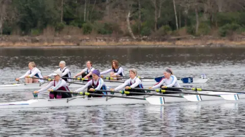 Clive Penkett Two crews of four women sit in boats waiting to start racing. They are all leaning forward holding their oars with their knees bent. Behind them you can see the shores of Derwentwater  