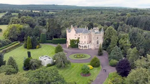 Pink castle nestled among trees and lawn in long short taken from sky