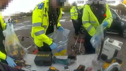 Police officers in high-visibility jackets putting tools in plastic evidence bags.