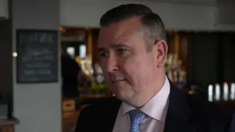 A man in a suit and tie is standing in a bar, with bottles, glasses and chalkboard signage visible behind him.