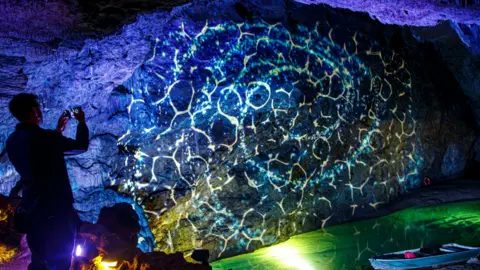 Wookey Hole A bluish light display projected onto a huge rock in a cave with a visitor to the attraction taking a photo in the foreground. 