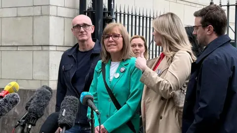 Lyra McKee's sister Nichola is wearing a green jacket and is speaking into a microphone outside court. She has sandy coloured hair and glasses. Other family members stand beside her