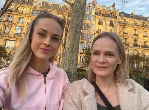 Vicky Vicky, a woman with long blonde hair and wearing a pink top, stands next to her mother Jo, who is also blonde and wearing a beige coat.