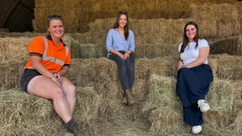 Ben Schofield/BBC Three women sitting on hay bales and looking directly to the camera. Bonnie Oakley is sat on the left of frame. She is wearing dark shorts and a bright orange, high visibility T-shirt. She is smiling and leaning slightly towards the centre of the picture. She has a pair of sunglasses on her head and her dark blonde hair is pulled back behind her head. In the middle of the picture is Alice Northern, who is also smiling. She is wearing a loose blue shirt, jeans and boots. Her hands are placed in her lap. She has brown, straight hair worn down and falling over her shoulders. She also has a pair of sunglasses on her head.
