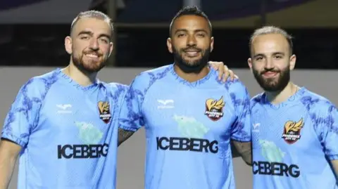 Bader Al-Akkad Amer Al-Akkad, Danny Simpson and Bader Al-Akkad stand together in light-blue Phoenix United shirts on an astro football pitch, with Simpson having a football in front of his feet. 