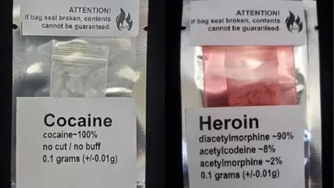 A photo of two labelled batches of drugs sold by Kalicum and Nyx. One package is labelled '0.1 grams of Cocaine' while another is labelled '0.1 grams of Heroin.' 