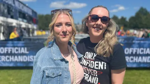 Two sisters stand inside the entrance to TRNSMT at Glasgow Green - on ein a denim jacket and white skirt, the other in a red skirt and black t-shirt