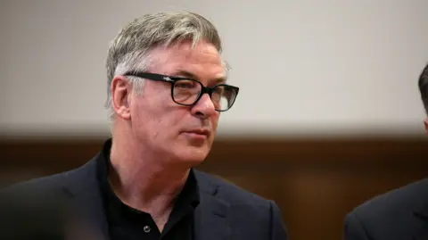 Reuters Alec Baldwin wears a grey suit and black glasses.