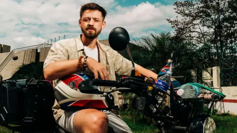 Harry Jackson sits on a moped. He has short dark hair and a beard and is wearing a T-shirt, cream-coloured top, a necklace and shorts, He is by a building with foliage all around him.