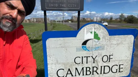 Bhupinder Sandhu Bhupinder Sandhu taking a selfie with a road sign that says "city of Cambridge". He is to the left of the photo, wearing a red rain jacket and a black turban. 