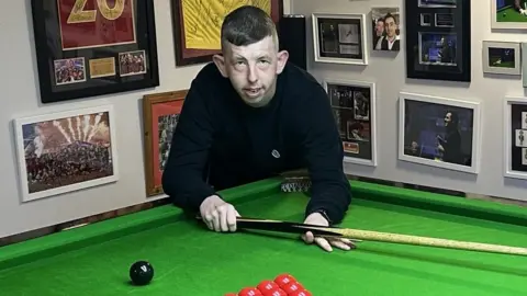 David Church holding a cue and leaning on a snooker table. He is surrounded by snooker memorabilia