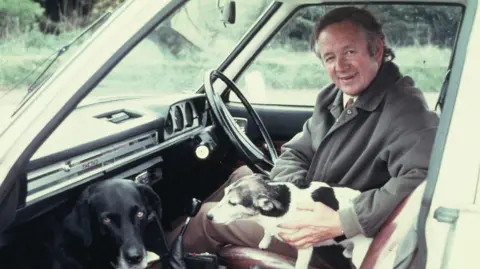 A man sits in the driver's seat of an old-fashioned car with two dogs.