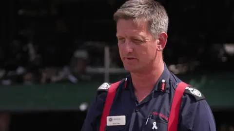 Andy Cole stands giving an interview. He wears a uniform consisting of a navy blue collared shirt, with black patches on the sleeves and red brace straps on his shoulders. He has short greying hair and is looking slightly off camera. 