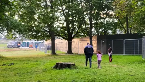 Save Gunnersbury Park Two children and man walk through the park near temporary metal fences in August 2023