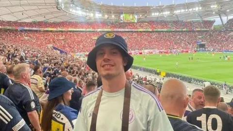 Daris Nesbitt A smiling Daris, who is wearing a blue Scotland bucket hat and a light blue Scotland shirt, poses for a picture in the stands of a packed stadium. 