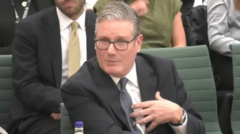 Sir Keir Starmer gives evidence at the Liaison Select Committee, wearing a black suit, a white shirt and a grey tie. 