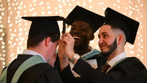NMITE Two men helping another man put his graduation cap on. One man is fitting the cap and the other is laughing in the background. They are all wearing black gowns with sage green hoods.