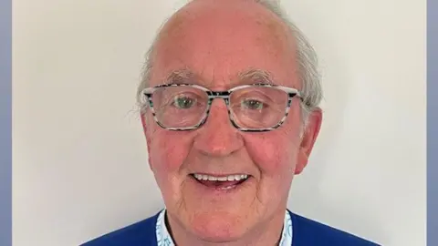 John Gerard Boyle who has short white hair and is wearing black and white framed glasses and a blue jumper over a white shirt. He is smiling broadly. 