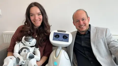 Dr Ioanna Giorgi and Dr Giovanni Masala sat on a sofa with two small child-like robots, with NAO and Buddy