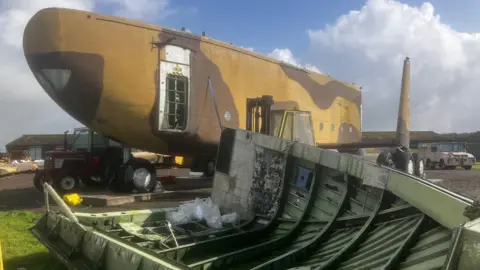 BBC Parts of the aeroplane are laid out on a field. It is painted in shades of brown and green.