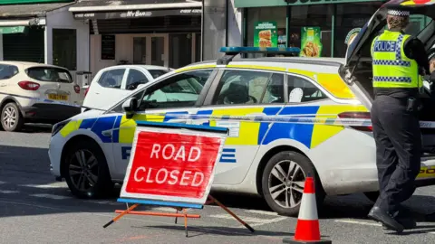 Police car and officer with police tape and road closed sign outside parade of shops.