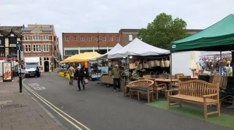 Darren Rozier/BBC Market stalls are set up on a street in a town. They are selling baskets, furniture and fruit and vegetables. Two shoppers are crossing the road to and from the stalls. There is a white van parked on the side of the road nearby and redbrick buildings in the background