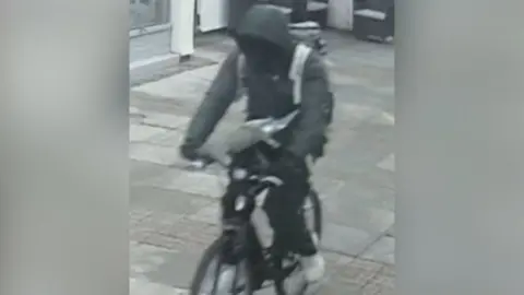 A CCTV still of a person riding a black bike along pavement. They are wearing a grey coat with its hood up.