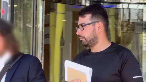 BBC Andrew Manuel Carvalho Franco walks out of a modern building with glass doors and metal framing. He has short dark hair and black-framed glasses, and is wearing a black T-shirt while holding pieces of paper.