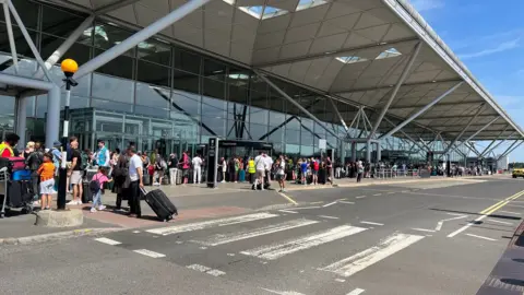 The outside of the departures area at Stansted Airport, with groups and queues of people outside the terminal building beside the dropping off bays on the road