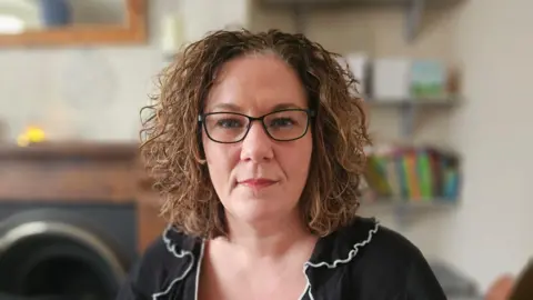 BBC Gemma Whysall is wearing black glasses and has shoulder length curly hair. She is wearing a black blouse with a white trimmed frilly collar. She is looking directly at the camera and there is a book case and a fireplace behind her.